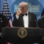 Donald Trump in a tux points to his forehead during a press briefing. He's flanked by Kash Patel and Todd Blanche.