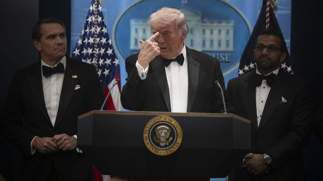 Donald Trump in a tux points to his forehead during a press briefing. He's flanked by Kash Patel and Todd Blanche.