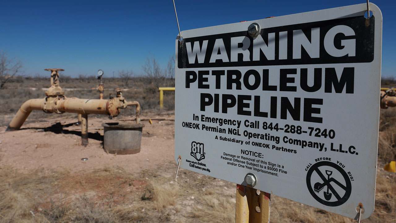 Yellow oil pipelines in a barren West Texas landscape. In the foreground, a sign reading "Warning Petroleum Pipeline"