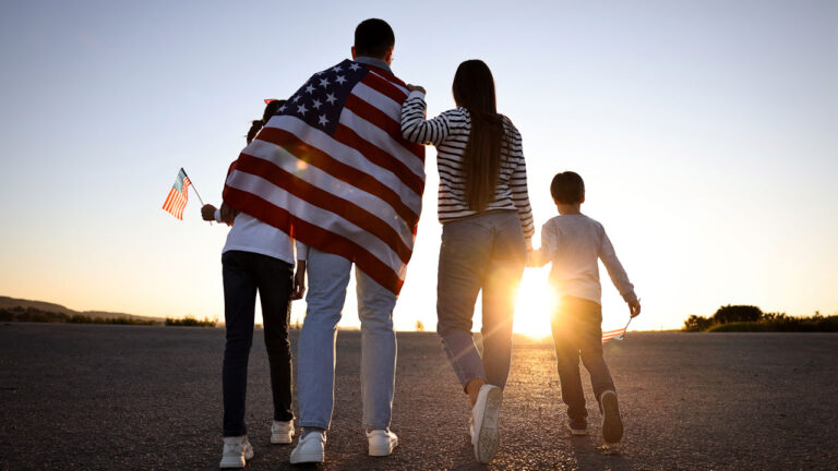 A mother and father walk with their two kids toward a sunset, a flag draped on the father's back and a flag in the hand of the taller sibling.
