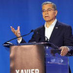 Xavier Becerra behind a podium speaks, with a hand raised.