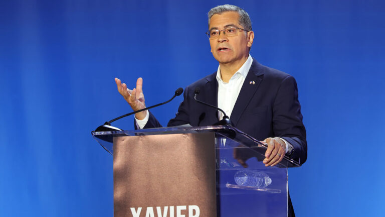 Xavier Becerra behind a podium speaks, with a hand raised.