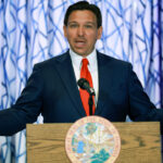 Ron DeSantis behind a podium with his mouth open and arms stretched wide.