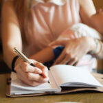 A mother writes in a notebook while holding her sleeping child at a table.