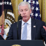 Energy Secretary Chris Wright holds his hands up while speaking behind a podium with flags behind him.