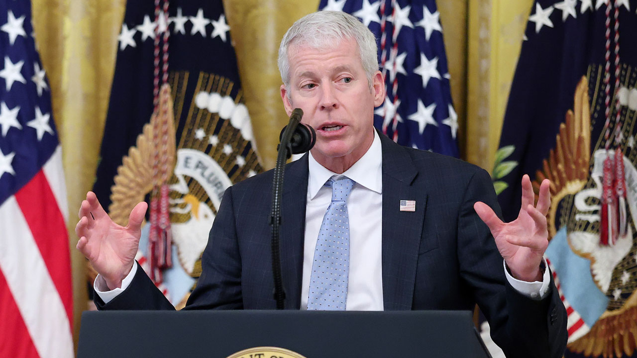 Energy Secretary Chris Wright holds his hands up while speaking behind a podium with flags behind him.