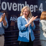 Abigail Spanberger, flanked by Jay Jones and Ghazala Hashmi clap in front of bus during campaign rally.