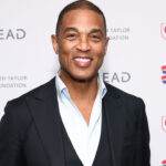 Former CNN host Don Lemon in black jacket and open white shirt stands against a white background at Elizabeth Taylor AIDS Foundation event.