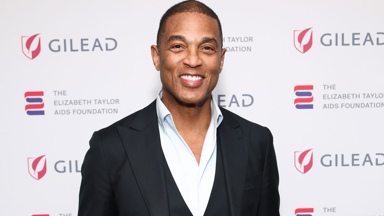 Former CNN host Don Lemon in black jacket and open white shirt stands against a white background at Elizabeth Taylor AIDS Foundation event.