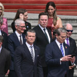 Pete Hegseth and Scott Bessent surrounded by business leaders including Elon Musk and Tim Cook stand on steps.