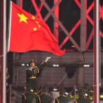 Chinese soldier points to large Chinese flag as row of Chinese soldiers salute.