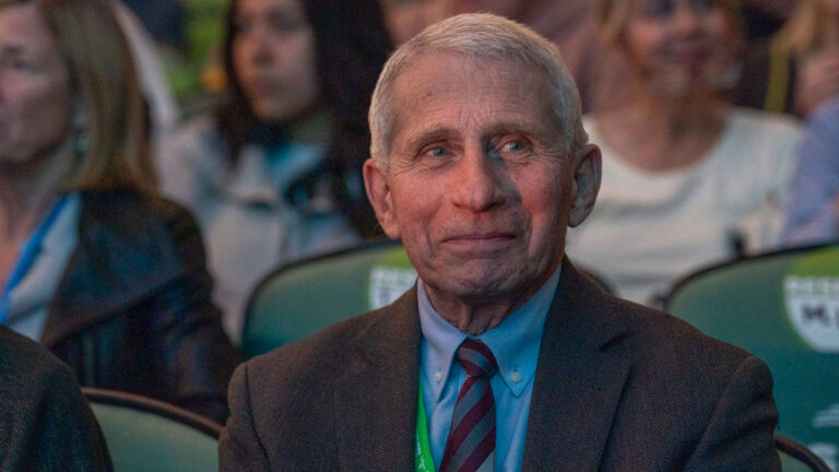 Dr. Anthony Fauci in a grey suit and blue shirt grins while seated.