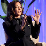 A seated Kamala Harris raises her hands as she makes a point during a talk.