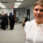 Aimee Bock, in a white blouse, stands in a room with others in the background.