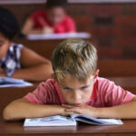Bored, restless boy rests head on arms as he sits at his classroom desk, while female students behind him are working away.