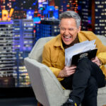 Greg Gutfeld sits in a gray armchair, laughing while holding papers and a clipboard. Behind him is a cityscape backdrop with the word GUTFELD! in bright lights.