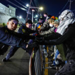 Masked protesters push against police barricades as an officer looks into the camera.