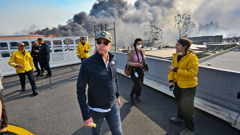 Gavin Newsom, in cap and sunglasses walks as black smoke billows behind him.