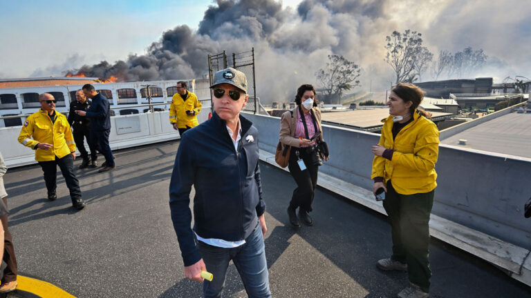 Gavin Newsom, in cap and sunglasses walks as black smoke billows behind him.