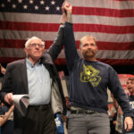 Bernie Sanders lifts Graham Platner's arm at a rally, with American flag as a backdrop.