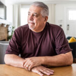 Middle-aged Hispanic man in a brown shirt sitting at a dining room table looking out toward a window.