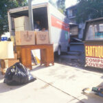A moving van in a street awaiting furniture to be loaded, a bag of trash and a newspaper vending machine with banner "Earthquake coverage."