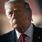 A man with light hair wearing a dark suit, white shirt, and red tie looks ahead with a serious expression. An American flag pin is visible on his jacket lapel, hinting at his involvement in Iran-related affairs. The background is dark and out of focus.