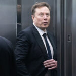 Elon Musk in a dark suit looks toward camera.