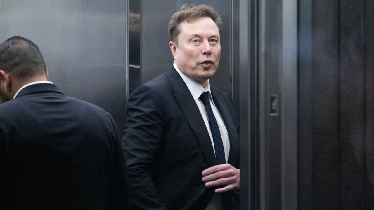 Elon Musk in a dark suit looks toward camera.