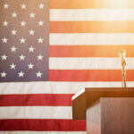 A podium in front of an American flag. Bright light hangs above the microphone.