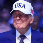 A grinning Donald Trump in a blue suit with white baseball cap with USA in blue lettering.