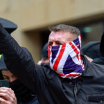 A protester, his face covered in a UK flag, raises his fist as other protesters and a police officer stand behind him.