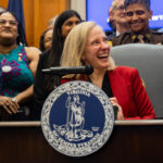 A laughing Abigail Spanberger sits at a desk surrounded by smiling, laughing people.