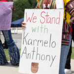 Protester in African garb holds sign reading "We Stand With Karmelo Anthony."