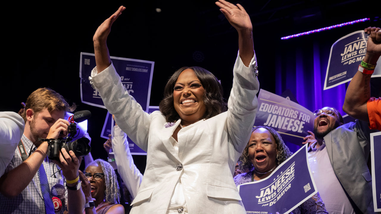 Janeese Lewis George in a white suit waves with both hands in the air as supporters celebrate behind her.