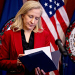 Abigail Spanberger in a red jacket looks down at a file. Behind her, two American flags and a Virginia state flag.