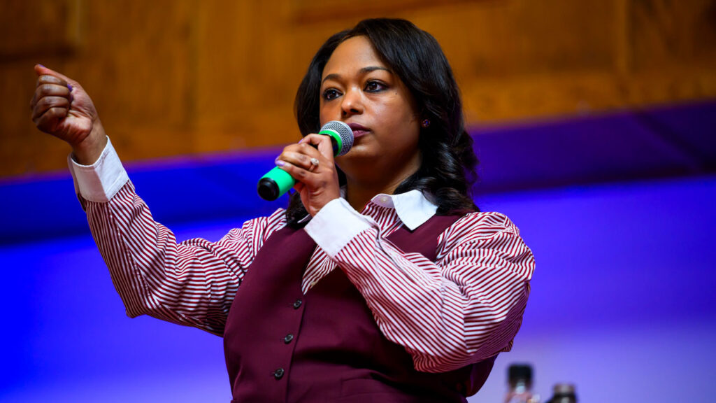 Janeese Lewis George in a maroon vest and maroon and white striped shirt holds a microphone to her mouth and has her right hand stretched out and clenched.