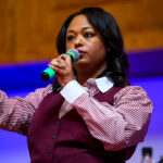 Janeese Lewis George in a maroon vest and maroon and white striped shirt holds a microphone to her mouth and has her right hand stretched out and clenched.