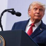 A grinning Donald Trump leans to his left while at a podium featuring the presidential seal.