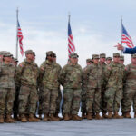Secretary of War Pete Hegseth points as he's standing with and addressing National Guard troops with numerous American flags behind them.