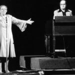 Black-and-white photo of singer Kate Smith on stage with her arms spread wide. To her left a man on keyboards.