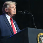 Side view of Donald Trump at podium against a dark background with his right arm gesturing to the side.