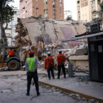 Several workers stand in front of the rubble of a collapsed building as a bulldozer works with the rubble.