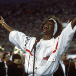 Whitney Houston in a white, with blue and red trim pullover, has her arms outstretched as she concludes singing "The Star-Spangled Banner" during Super Bowl XXV