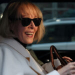 Close-up of E. Jean Carroll in sunglasses and white coat smiling while seated in car.
