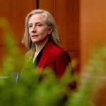 Gov. Abigail Spanberger in red jacket, seen through a row of marijuana plants in the foreground.