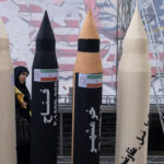 An Iranian woman walks behind mock-ups of four Iranian missiles against a backdrop of an drawing of a scowling Donald Trump.