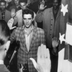 Elvis Presley stands with a group of young men at an induction center, raising their right hands as they are sworn into the United States Army by an officer standing next to an American flag.
