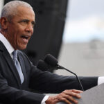 Barack Obama at a podium speaking with arm outstretched.