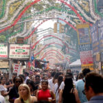 A crowded Italian street festival with food stalls, colorful hanging decorations, and many people walking and enjoying the event on a sunny day.